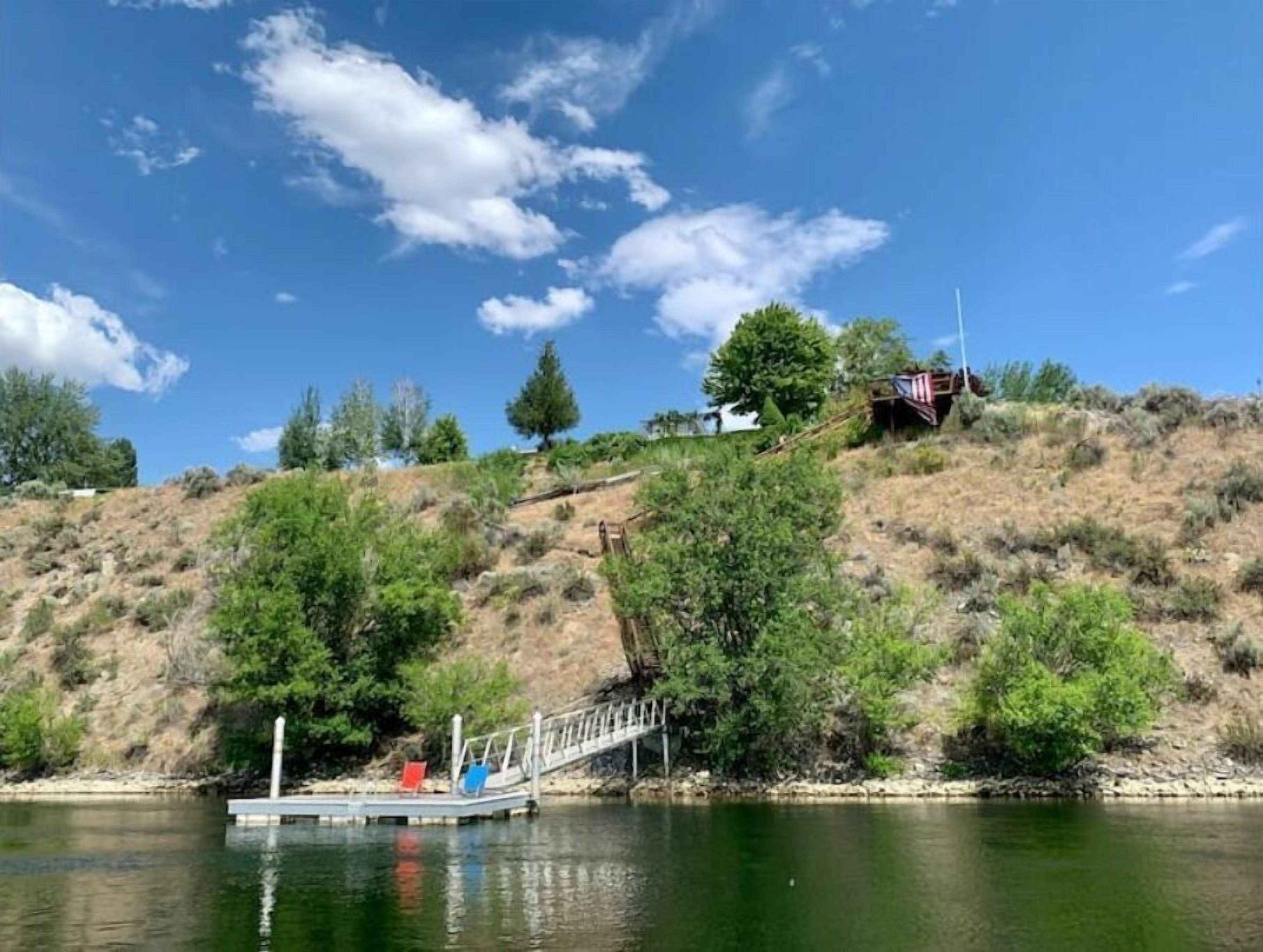 A dock extending into a calm body of water, surrounded by greenery and a hill with sparse vegetation.