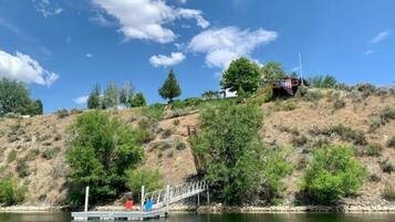 A dock extending into a calm body of water, surrounded by greenery and a hill with sparse vegetation.