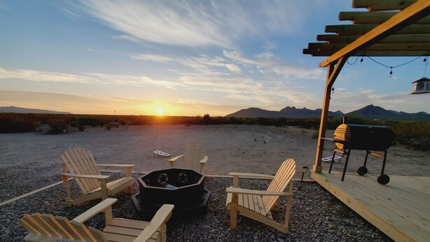Une terrasse dotée de chaises en bois et d’un foyer, offrant une vue sur un paysage désertique avec des montagnes à l’horizon.