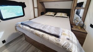 Une chambre compacte dans un camping-car, comprenant un lit, une fenêtre, un miroir et une petite table avec un ventilateur.
