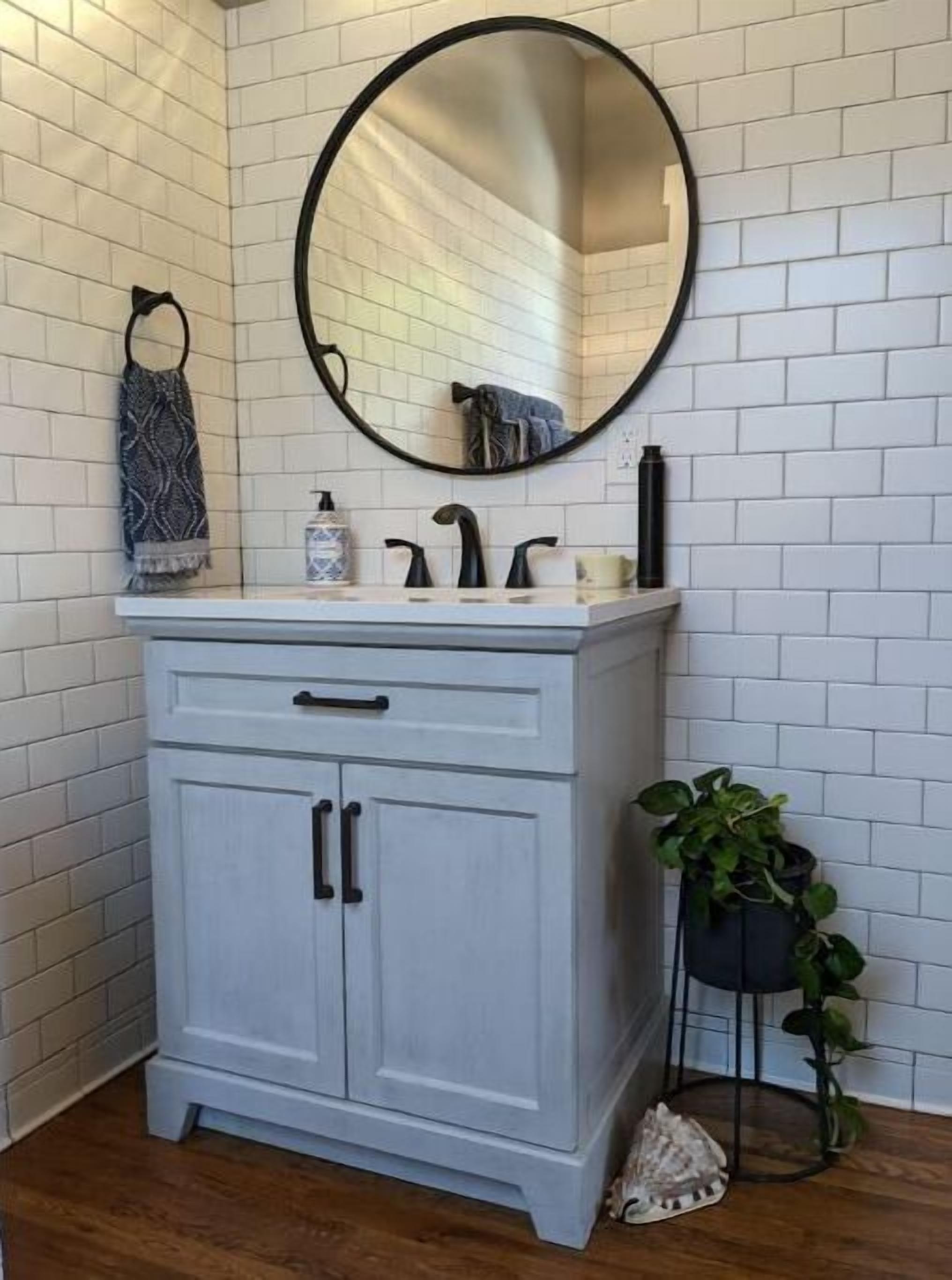 Een badkamer met een witte betegelde muur, een witte wastafel met twee deuren, een ronde spiegel, een handdoekhouder en een potplant.