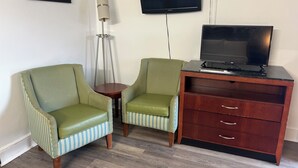 Una habitación con dos sillones tapizados en verde, una pequeña mesa redonda, una lámpara de pie alta, un armario de madera con cajones y un televisor de pantalla plana montado en la pared.