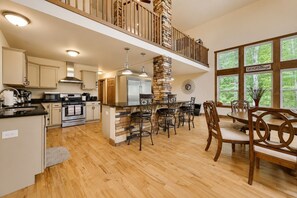 A spacious kitchen with a central island, wooden flooring, and a dining area with a high ceiling and natural light.
