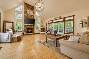 A spacious living room with a fireplace, large windows, and a high ceiling.
