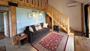 A spacious living area with a wooden staircase, a dark sofa with cushions, a red patterned rug, and a small wooden table. - Prince Edward Island 2 bedroom waterfront cottage in South Rustico (New Glasgow)