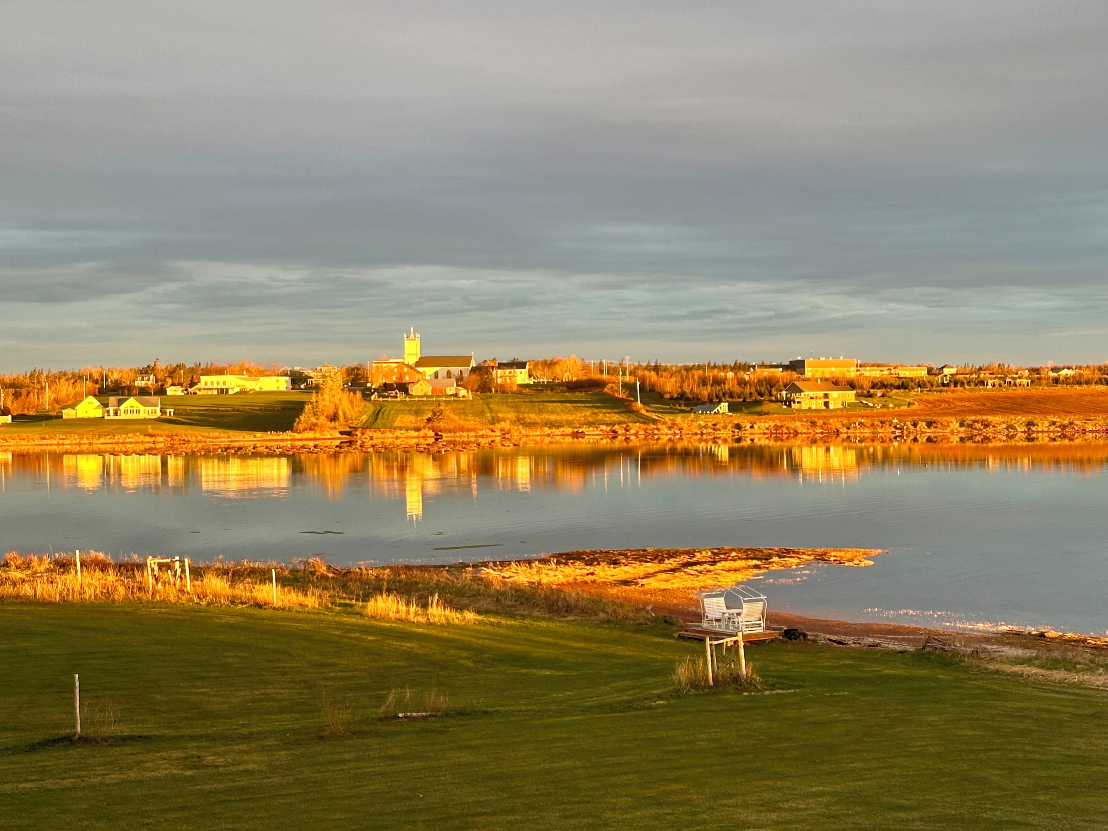 A lakeside view with a church, houses, and a grassy area with a bench.