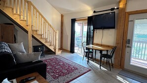 A spacious living area with a wooden staircase, a flat-screen TV mounted on the wall, a dining table with chairs, and a red patterned rug. - Prince Edward Island 2 bedroom waterfront cottage in South Rustico (New Glasgow)