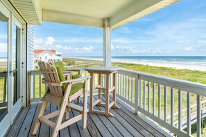 Eine Holzterrasse mit einer Bank und einem Tisch, mit Blick auf einen Strand und ein Haus im Hintergrund.