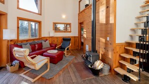A cozy cabin interior with a wooden floor, a red sofa, a wooden rocking chair, a fireplace, and a wooden staircase.