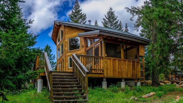 A wooden cabin with a staircase leading to a deck, surrounded by trees and rocks.