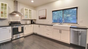 A modern kitchen with white cabinetry, a stainless steel range hood, a built-in oven, a dishwasher, and a window with blinds. - The Charming Home with a Pool (Yuba City)