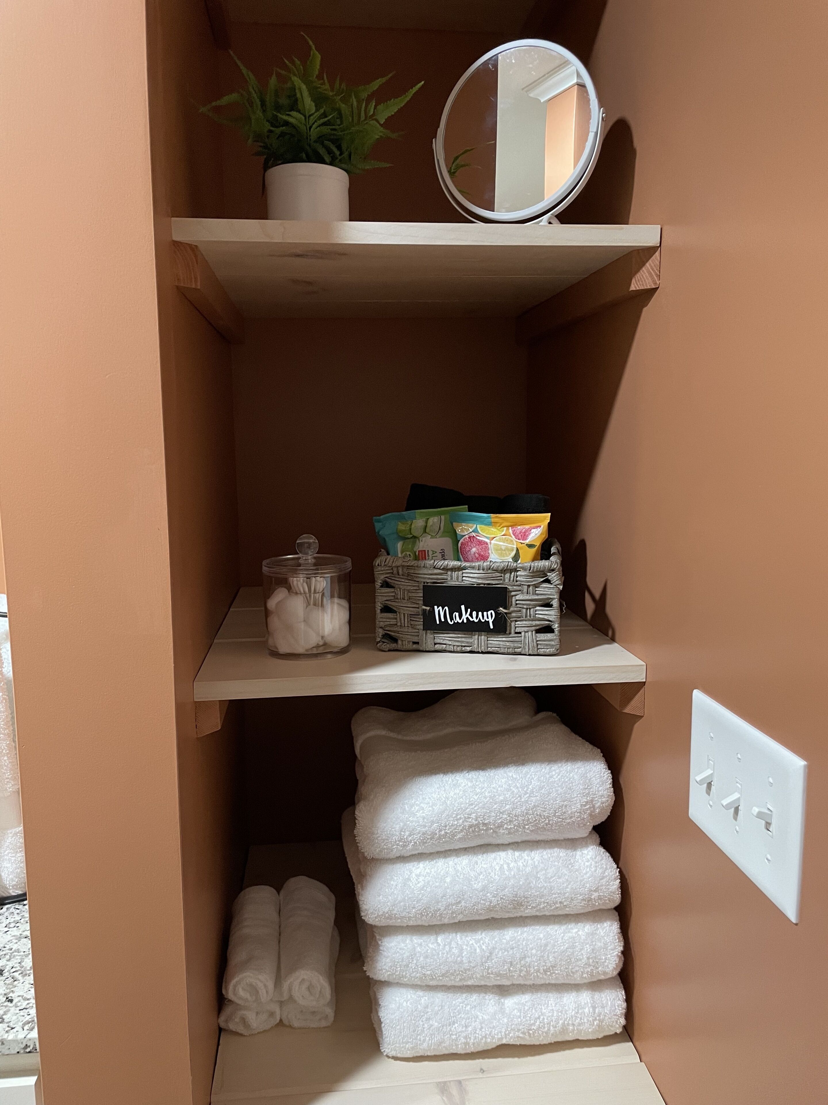 A bathroom with a shelf containing a potted plant, a round mirror, makeup items, and a stack of towels.