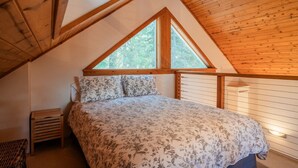 A bedroom with a sloped ceiling, wooden beams, a large window, a bed with floral bedding, and a bedside table. - Chinquapin Mountain Cabin w/ Jacuzzi and Wood Stove at Green Springs Inn (Ashland)