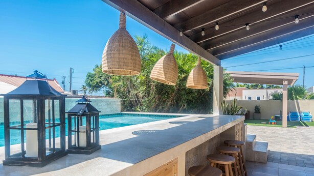 Un espace bar aménagé au bord de la piscine, doté d’un comptoir en béton, de tabourets en bois et de luminaires suspendus de style lanterne.