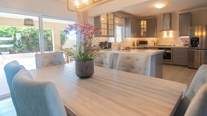 Une cuisine moderne équipée d’appareils électroménagers en acier inoxydable, un coin repas avec une table et des chaises en bois, et une vue sur un jardin grâce à de grandes fenêtres.