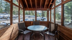 A wooden deck with a round table and chairs, large windows offering a view of a snowy landscape with trees.