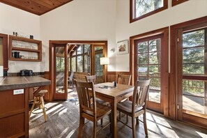 A wooden dining table with chairs, a kitchen area with shelves, and large windows.