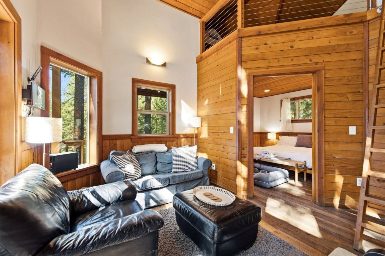 A cozy cabin interior with a sofa, ottoman, and a bed in the adjacent room.