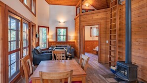 A cozy cabin interior with a dining area, a sofa set, a loft space, and a wood-burning stove.