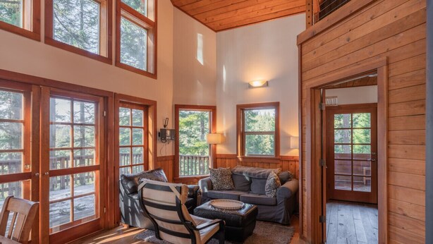 A cozy cabin interior with large windows, a wooden ceiling, and a seating area with a sofa and chairs.