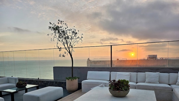 Eine Dachterrasse mit Blick auf den Sonnenuntergang über dem Meer, eine weiße Sofagarnitur, ein Couchtisch und ein Blumentopf.