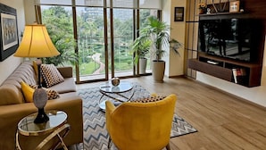 Ein modernes Wohnzimmer mit einer Couch, einem gelben Sessel und einem Fernseher an einer Holzwand. Große Fenster bieten Blick auf Grünflächen und Gebäude.