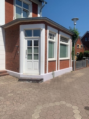 A corner house with a white door, two windows with shutters, and a small porch area.
