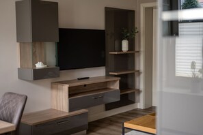 Ein modernes Wohnzimmer mit einem an der Wand befestigten Flachbildfernseher, Holzregalen und einem kleinen Tisch mit einem Stuhl.