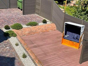 Una terraza de madera con una zona de estar integrada, rodeada por un muro de ladrillo y pequeños arbustos.