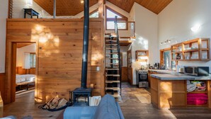 Ein gemütliches Blockhaus-Interieur mit Schlafzimmer im oberen Bereich, einer Holztreppe, einer Küchenzeile und einem Wohnraum mit Sofa und Kamin.
