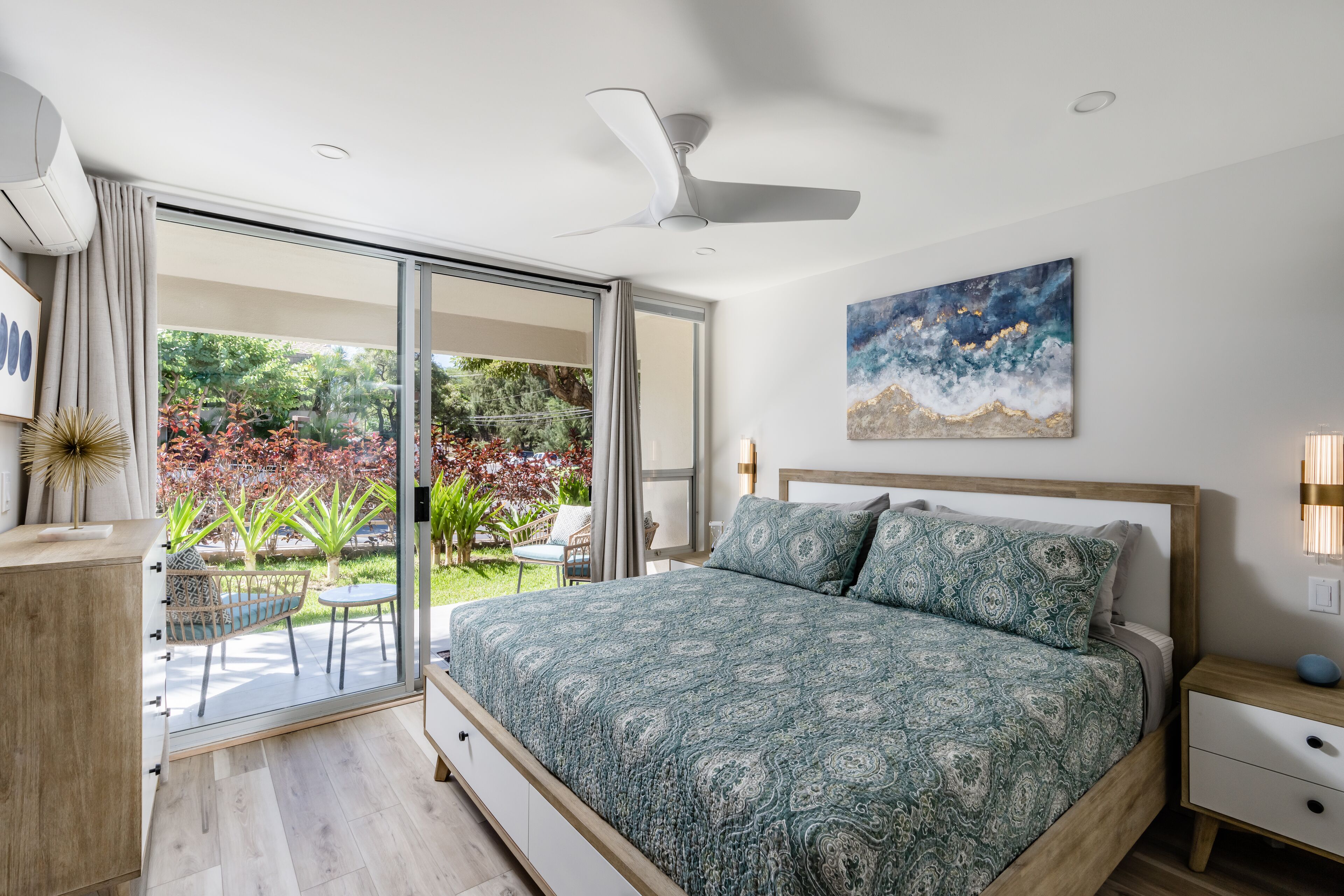 A bedroom with a large bed, a ceiling fan, a painting, and a view of a garden.