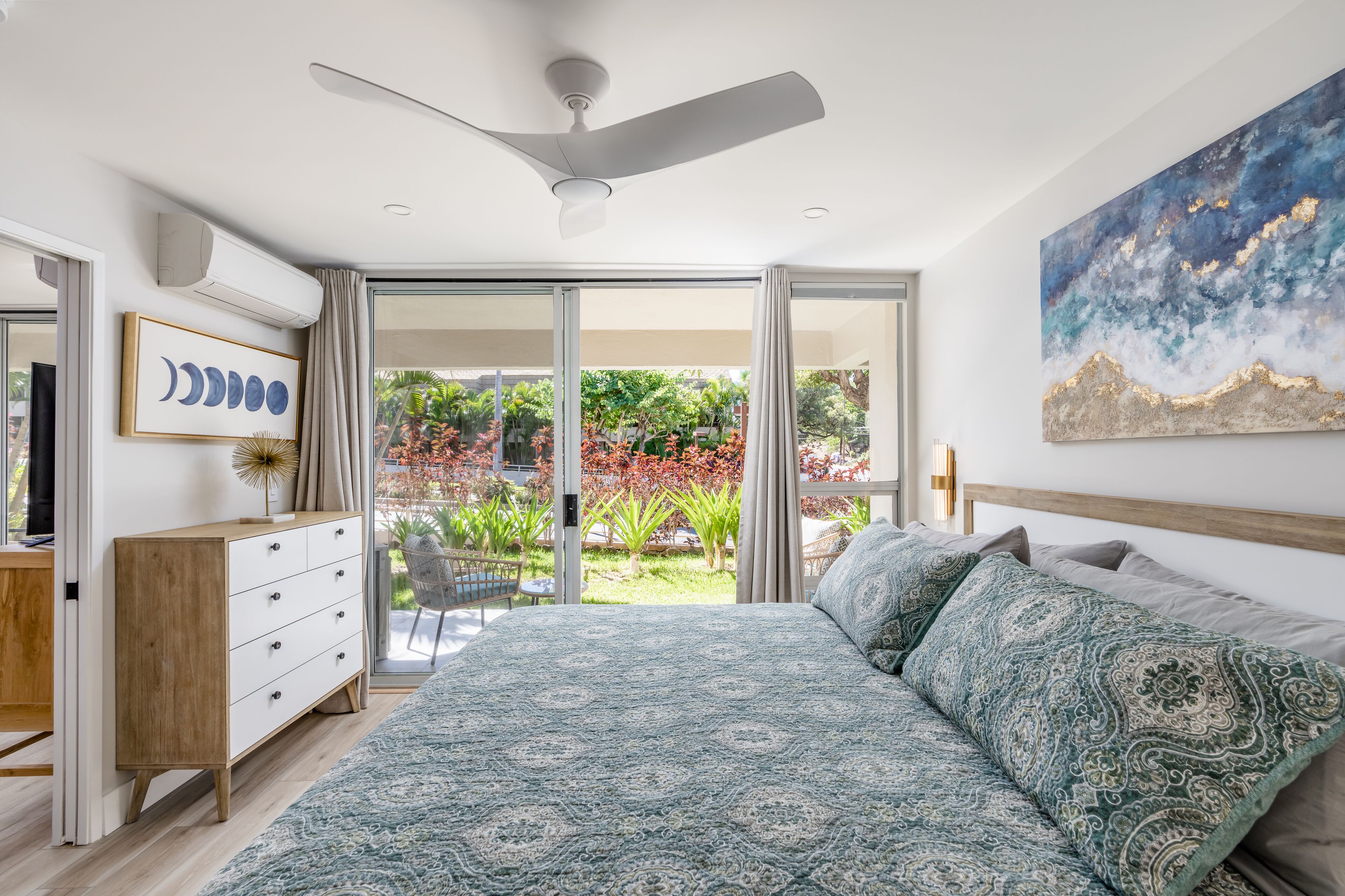 A bedroom with a bed, a ceiling fan, a dresser, and a large window with a view of greenery.