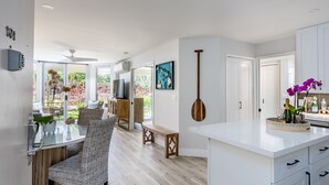 A modern kitchen with white cabinets, a dining area with wicker chairs, and a large window with a view of greenery.