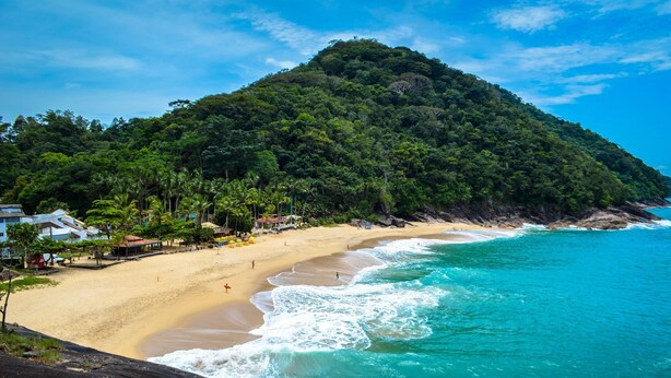 A beach with a sandy shore, clear turquoise waters, and a lush green hill in the background. - Severos mar 2 (Ubatuba)
