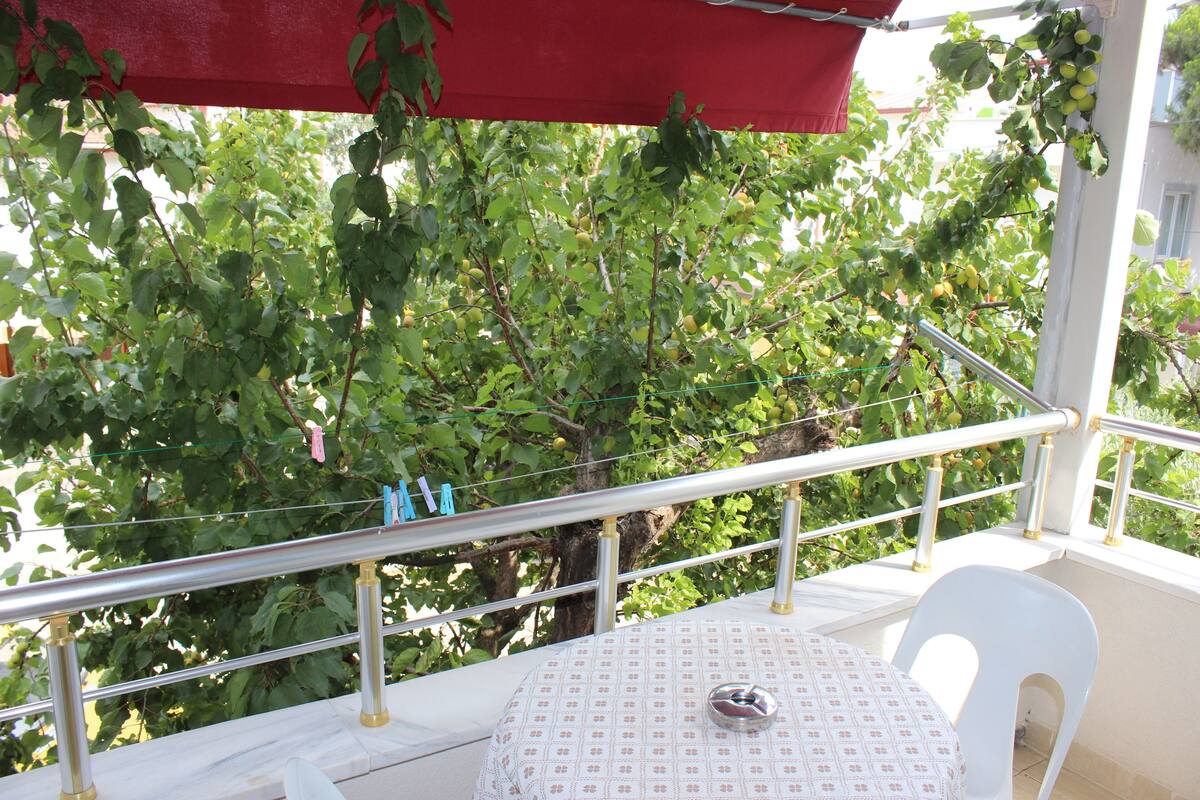 A balcony with a white table and chair, a red awning, and a view of green trees.
