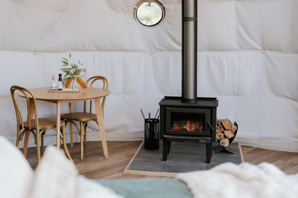 A cozy room with a round wooden table, two wicker chairs, a black wood-burning stove, and a small window.