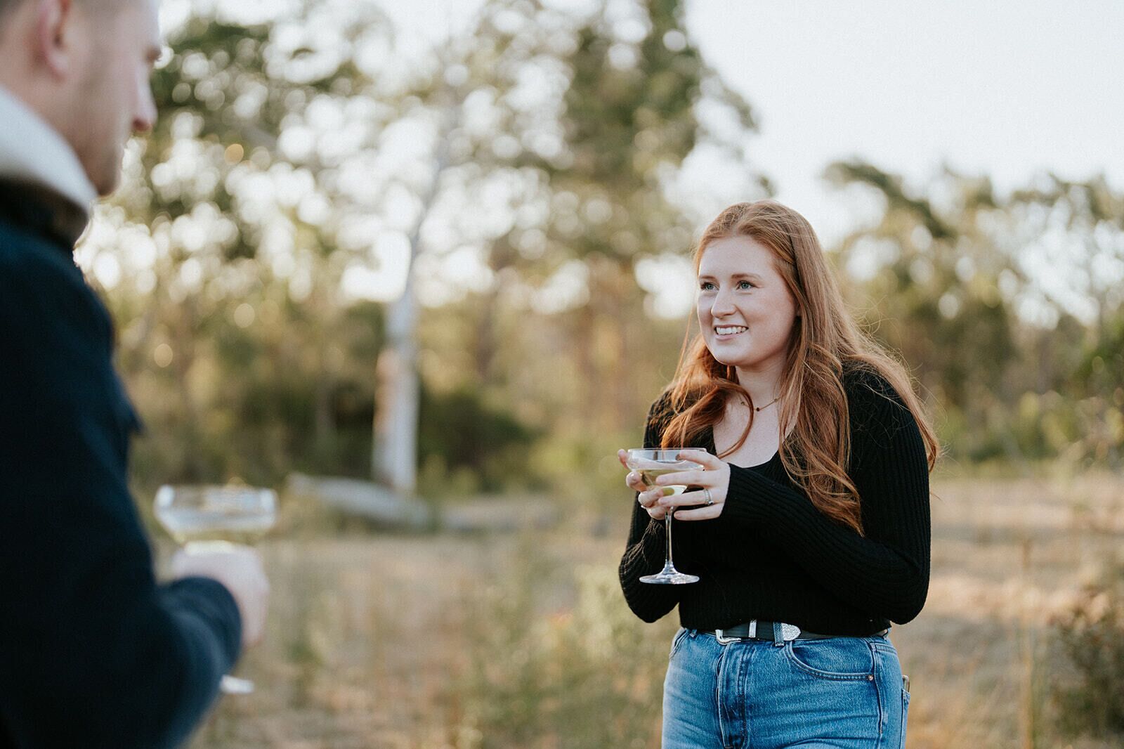 A person holding a glass, standing outdoors with trees in the background.