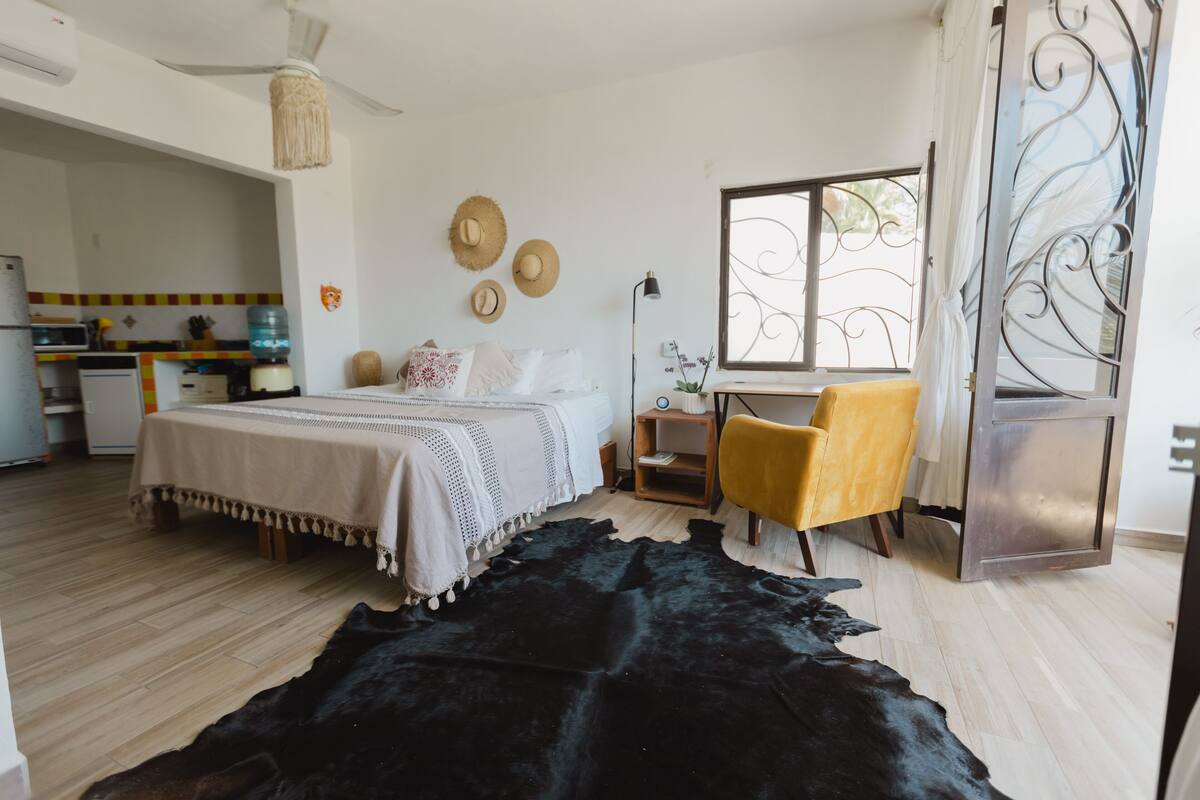 Un dormitorio con cama, escritorio, silla, ventana y una cocina al fondo.