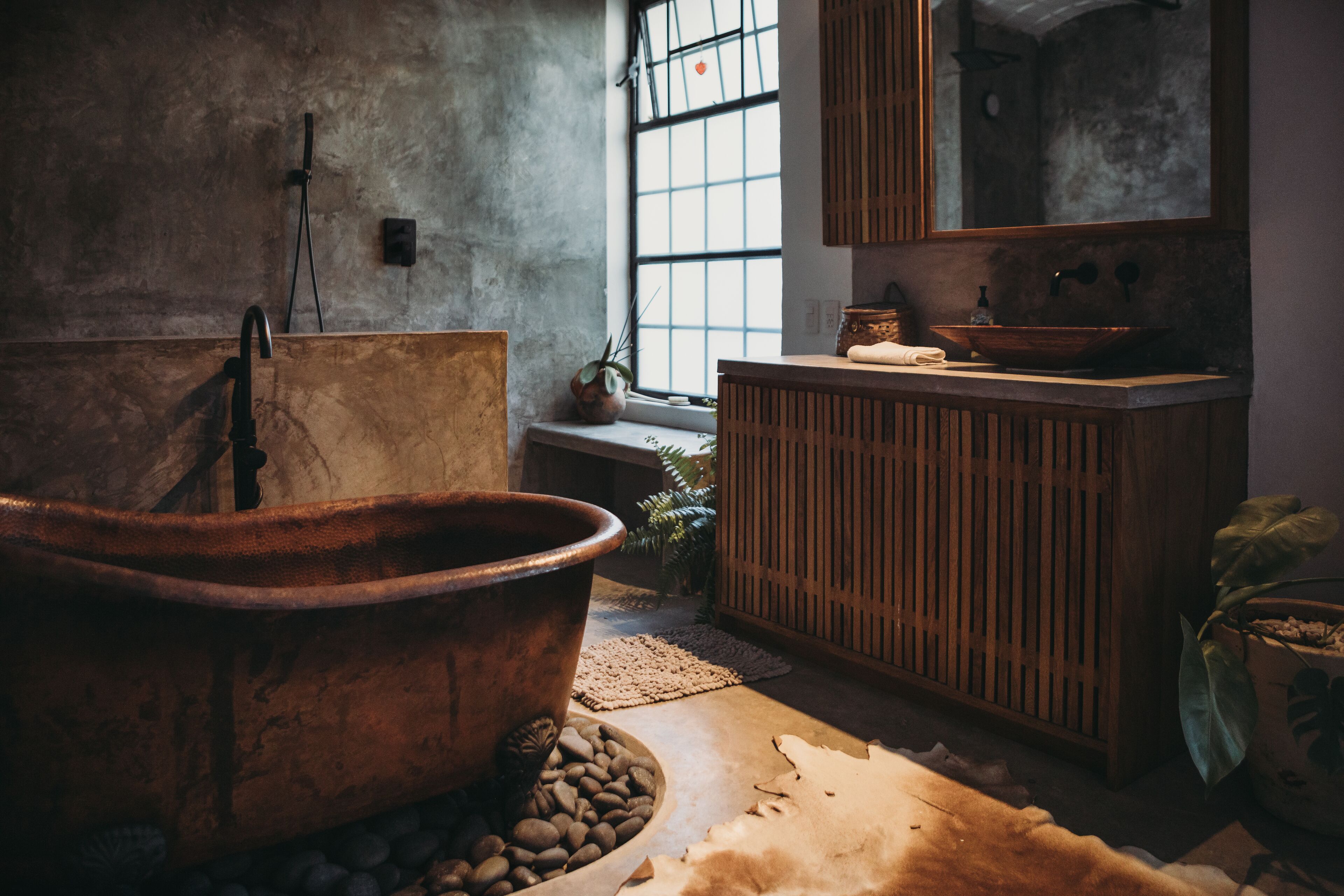 A rustic bathroom with a large copper tub, wooden vanity, and a large window providing natural light.