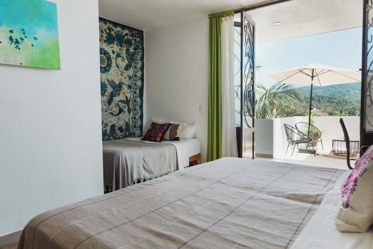 Un dormitorio con una cama grande, una mesita de noche pequeña, un tapiz colorido y un balcón con una silla y una sombrilla.