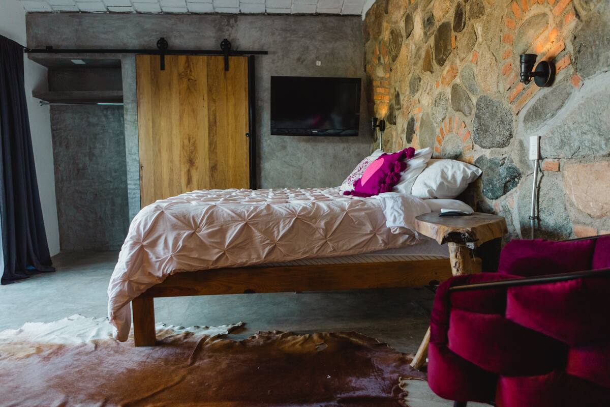 Un dormitorio con pared de piedra, una cama con estructura de madera, un televisor montado y una silla roja.