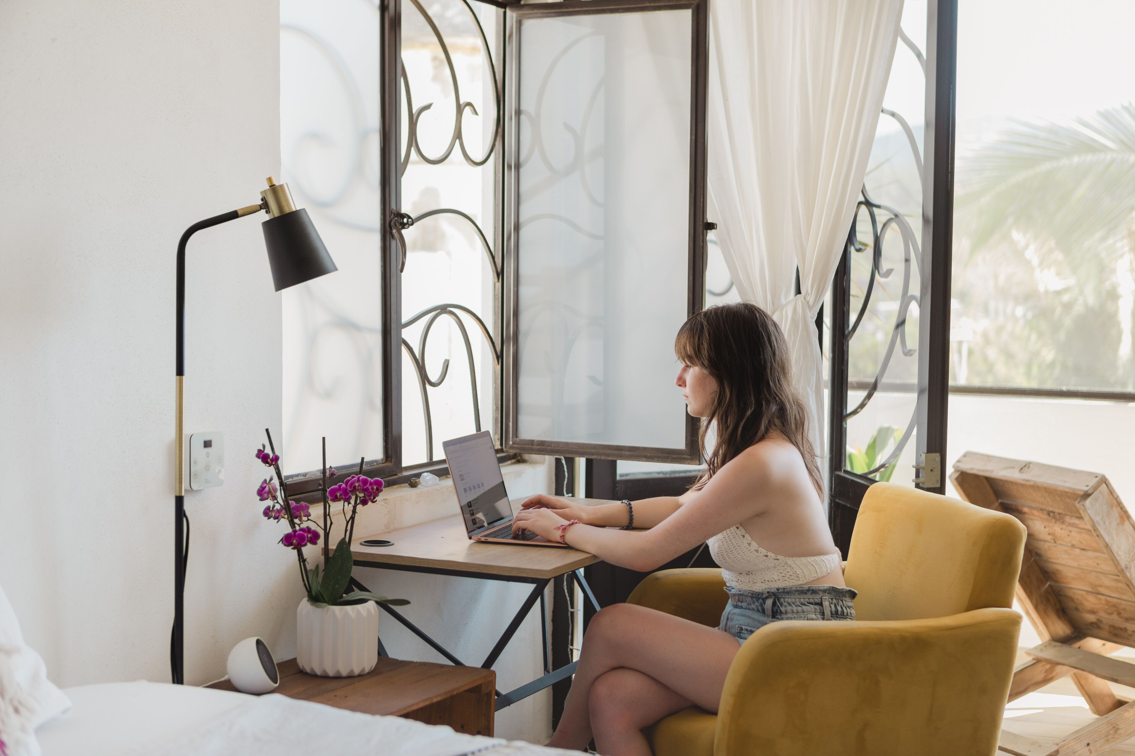 Una habitación con una cama, un escritorio con una computadora portátil, una orquídea en maceta y un ventanal con cortinas.