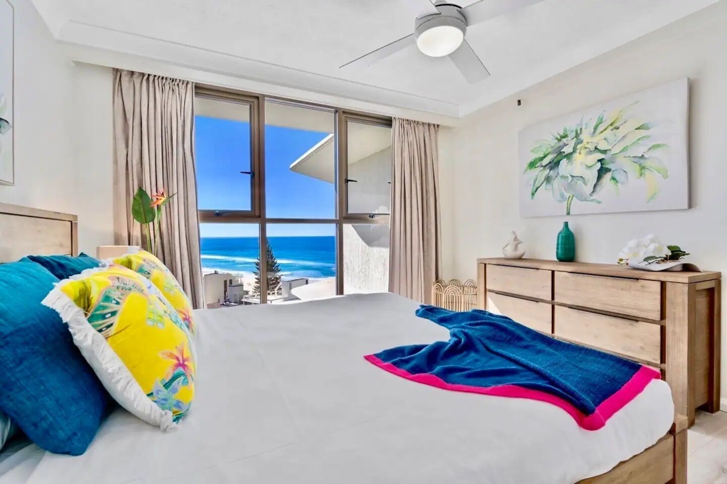 A bedroom with a large bed, a ceiling fan, a wooden dresser, and a painting.