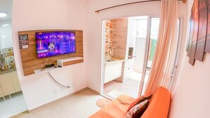 A modern living room with a flat-screen TV mounted on the wall, a sofa with cushions, and a sliding glass door leading to a balcony. - Geneis  (Ubatuba)