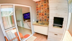 A modern kitchen with a built-in grill, a television, and a sliding glass door leading to a balcony with a wooden bench. - Geneis  (Ubatuba)