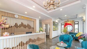 Un área de recepción con un mostrador de madera, un reloj y una zona de estar con sillones azul verdoso y una mesa de centro.