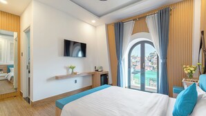Habitación de hotel moderna con una cama grande, un televisor montado en la pared, un escritorio pequeño con una lámpara y vistas a la ciudad a través de la ventana.