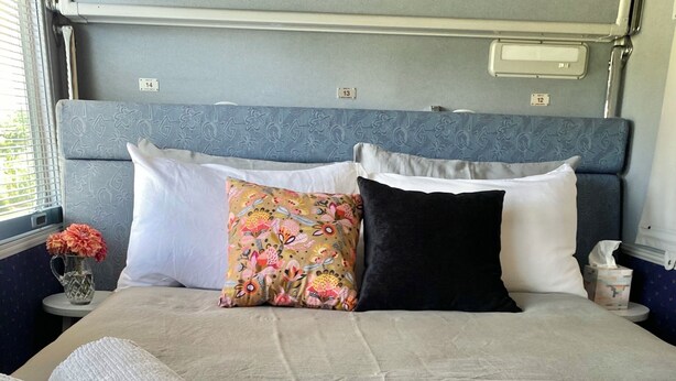 A neatly made bed with a grey bedspread, two rolled white towels, and a pink throw blanket.