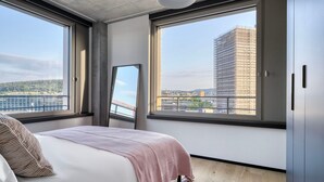 Ein modernes Schlafzimmer mit einem großen Bett, zwei Fenstern und Blick auf eine Stadtlandschaft.