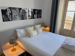 Une chambre d’hôtel avec un lit, des oreillers, une lampe de chevet, un mur orné de photos en noir et blanc et une fenêtre donnant sur un paysage.